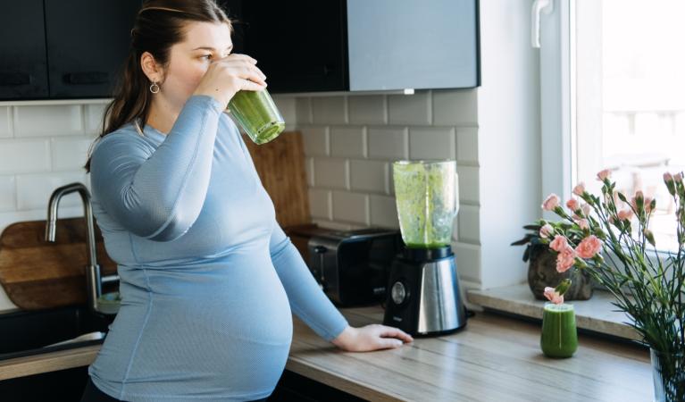A pregnant woman stands in the kitchen drinking a green smoothie.