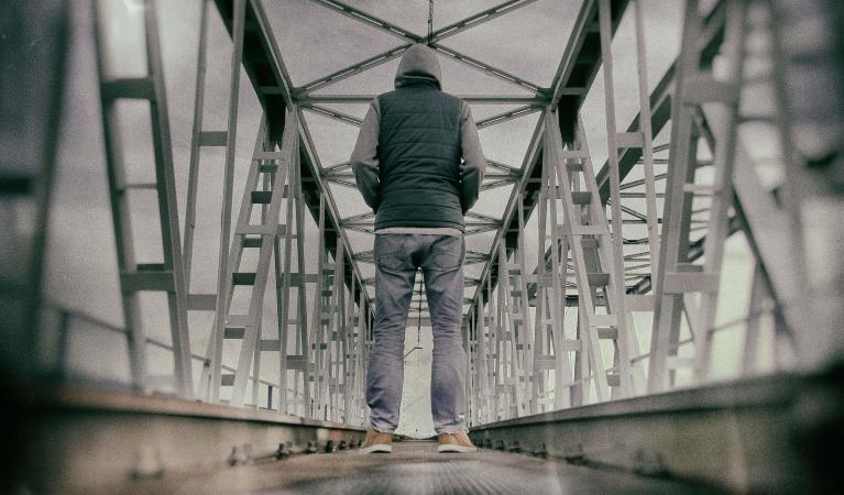 A person wearing a hooded jacket and jeans stands alone on a metal railway bridge, facing away from the camera.