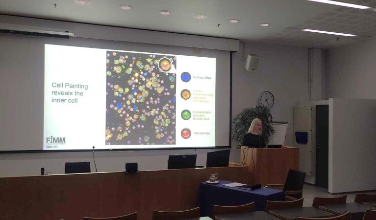 A doctoral researcher stands at a podium presenting her dissertation in a lecture hall, with a large screen displaying colorful cell painting images and labeled cellular components such as nucleus, mitochondria, and endoplasmic reticulum.