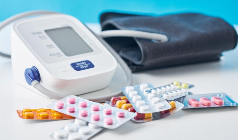 A digital blood pressure monitor and different medical pills on a white table.