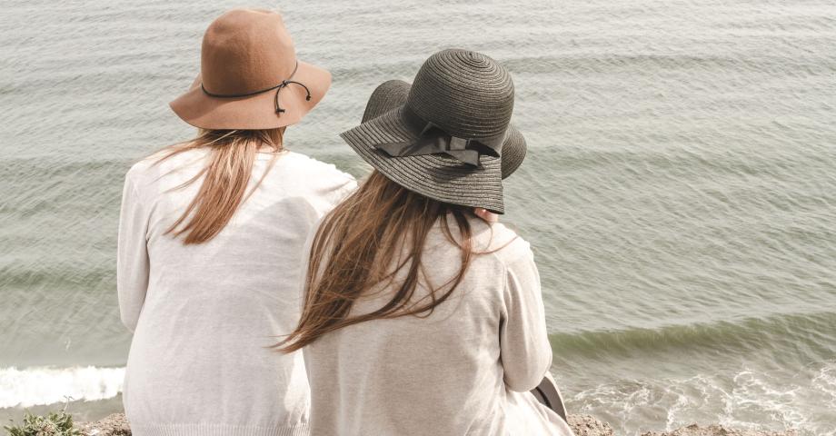 Kaksi naista kuvattuna selkäpuolelta istumassa rannalla aurinkohattu päässä.