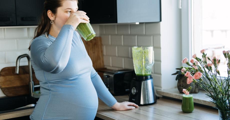 En gravid kvinna står i köket och dricker en grön smoothie. 