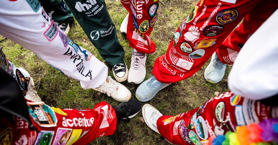 Eri värisiin opiskelijahaalareihin pukeutuneiden henkilöiden jalkoja ruohikolla.