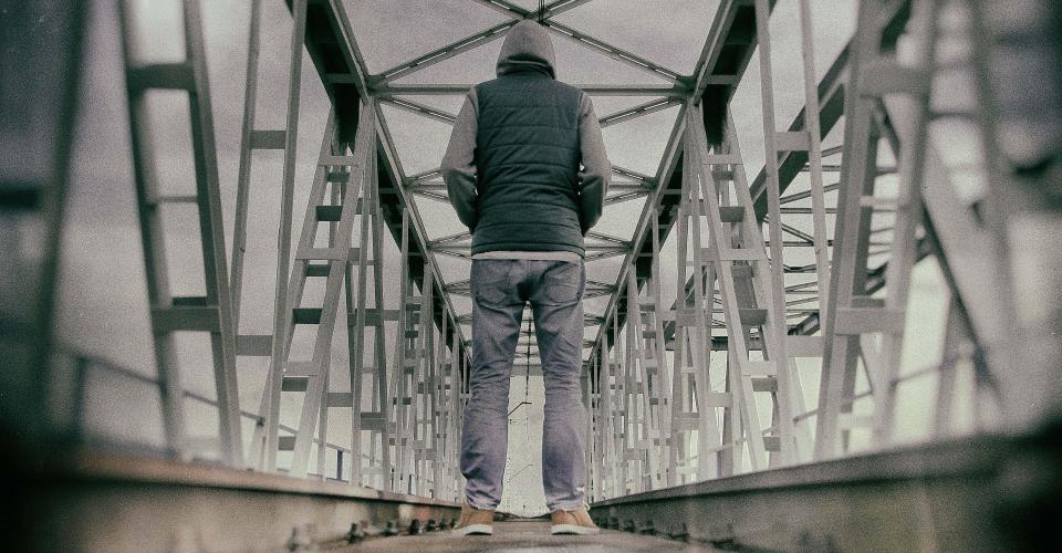 A person wearing a hooded jacket and jeans stands alone on a metal railway bridge, facing away from the camera.