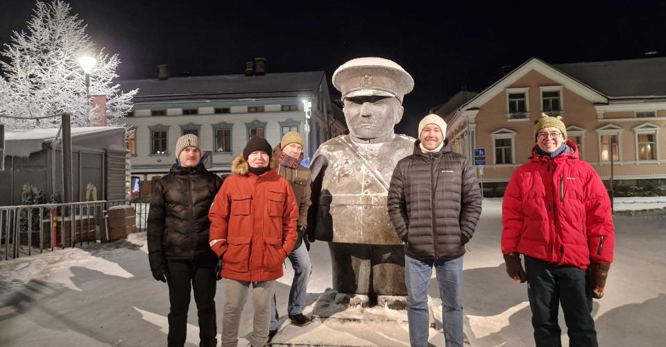Viisi talvivaatteisiin pukeutunutta henkilöä seisoo lumessa yöllä Oulussa jykevän patsaan ympärillä. Patsas ja maa ovat kevyesti lumen peitossa, ja taustalla näkyy valaistuja rakennuksia.