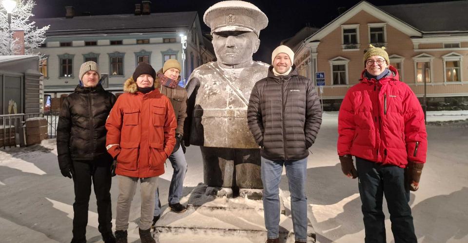 Five people in winter clothing stand in the snow at night in Oulu around a statue of a stout figure wearing a cap. The statue and ground are dusted with snow, and softly lit historic buildings line the background.