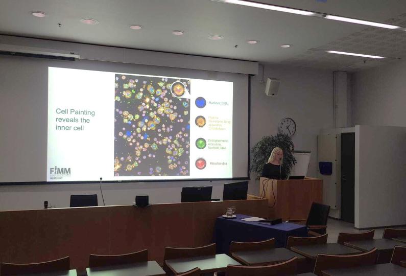 A doctoral researcher stands at a podium presenting her dissertation in a lecture hall, with a large screen displaying colorful cell painting images and labeled cellular components such as nucleus, mitochondria, and endoplasmic reticulum.