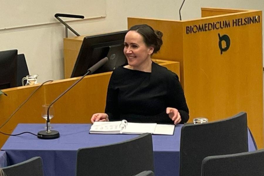 A woman in a black dress is seated at a desk in a lecture hall, smiling as she looks to her left.  A microphone, a folder and a glass of water are placed in front of her on the desk.