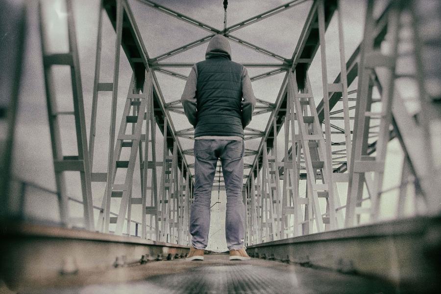 A person wearing a hooded jacket and jeans stands alone on a metal railway bridge, facing away from the camera.